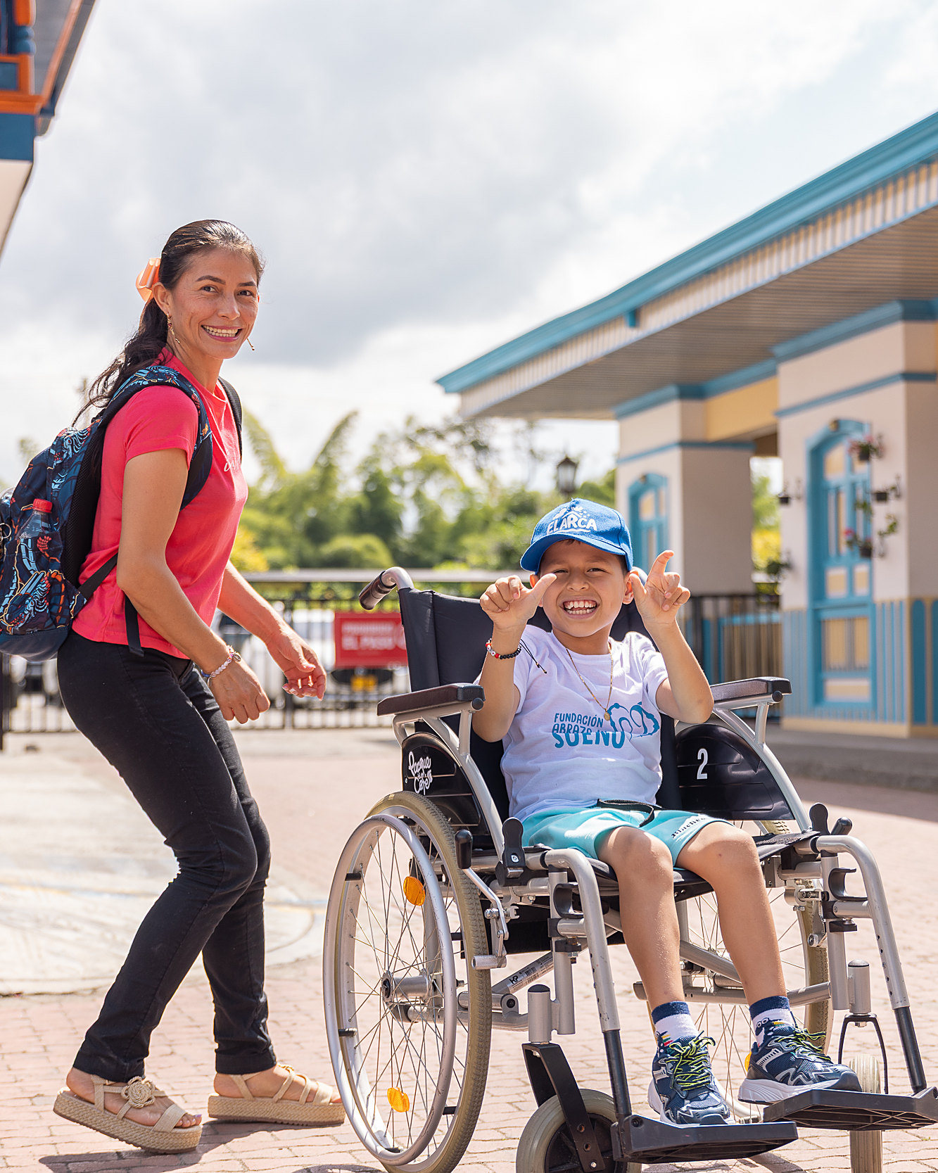 Un niño sonríe feliz en su silla de ruedas junto a una mujer de la Fundación, frente a una casa de balcones de colores del Eje Cafetero, Navidad 2025